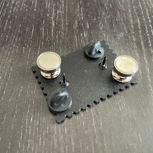 May include: The back of a black enamel pin with a silver metal pin back and a black rubber clutch. The pin is shaped like a rectangle with rounded corners and has a scalloped edge.