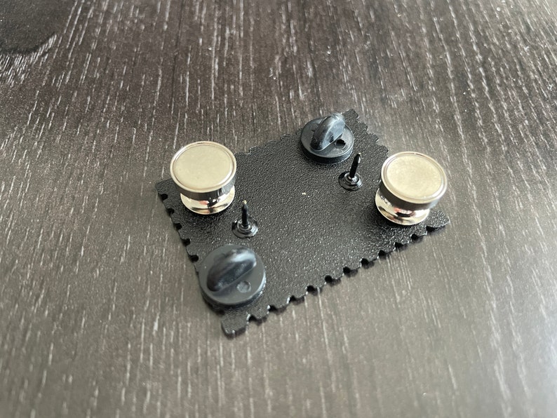 May include: The back of a black enamel pin with a silver metal pin back and a black rubber clutch. The pin is shaped like a rectangle with rounded corners and has a scalloped edge.