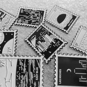 May include: A collection of black and white enamel pins featuring various cityscapes and text. The pins are shaped like postage stamps with a decorative border. The text on the pins includes "Gotham", "Neo-Tokyo", "Soul Society", "East Blue", "Las Noches", and "99&cent;", "92&cent;", "77&cent;", and "82&cent;".