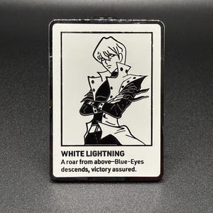 May include: A rectangular pin with a black and white illustration of a character. The pin has the text "WHITE LIGHTNING" and a quote about Blue-Eyes. The pin is set against a dark background.
