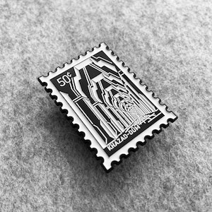 May include: A black and white enamel pin shaped like a postage stamp with the text "50¢" and "KHAZAD-DUM" and a graphic of a tunnel.