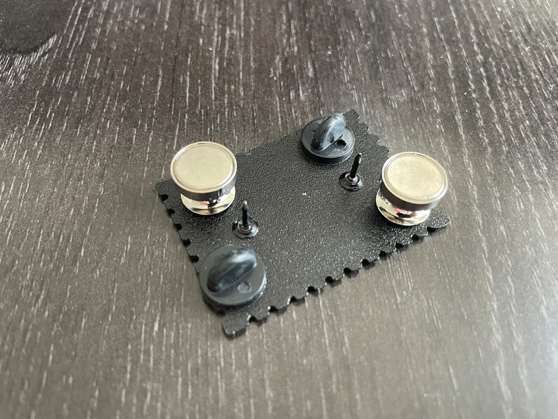 May include: The back of a black enamel pin with a silver metal pin back and a black rubber clutch. The pin is shaped like a rectangle with rounded corners and has a scalloped edge.