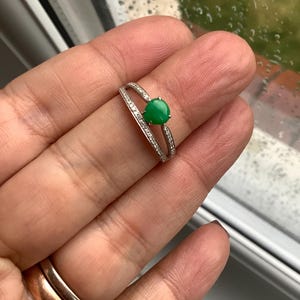 May include: A silver-toned ring featuring a small, round, vibrant green stone set in the center. The band is split into two thin, parallel arms, both of which are encrusted with tiny, clear, sparkling stones along their entire length.