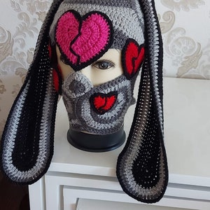 Puede incluir: Un gorro de punto gris y negro con largas orejeras. El gorro tiene una máscara de punto gris con aplicaciones de corazones rosas y rojos. El gorro está en una cabeza de maniquí blanca.