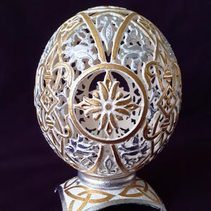 May include: Ornate, white, egg-shaped decorative object with gold and silver accents. The egg features intricate cut-out patterns and sits on a matching stand. The design includes floral and geometric elements, creating a detailed and elegant appearance.