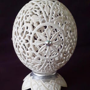 May include: Ivory-colored, intricately carved decorative egg. The egg features a delicate openwork design of floral and geometric patterns, with a central starburst design. It rests on a silver-colored base with a stylized floral motif. A small, iridescent gem is set in the center.