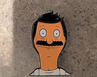 Könnte beinhalten: Eine schwarze und orangefarbene Emaille-Anstecknadel mit einer Comicfigur mit schwarzen Haaren, einem Schnurrbart und einem weißen Hemd. Die Figur hat einen überraschten Ausdruck mit großen, weißen Augen.