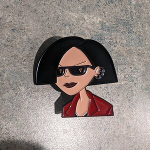 May include: Black and red enamel pin featuring a cartoon character with black hair, sunglasses, and a red jacket.