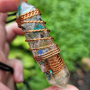 May include: A clear crystal pendant wrapped with copper wire. The crystal has blue and green inclusions and is suspended from a black cord. The copper wire is tightly wound around the crystal in a spiral pattern.