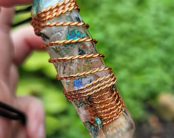 May include: A clear crystal pendant wrapped with copper wire. The crystal has blue and green inclusions and is suspended from a black cord. The copper wire is tightly wound around the crystal in a spiral pattern.