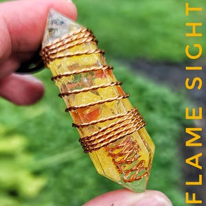 May include: A yellow crystal pendant wrapped in copper wire. The crystal is a light yellow color with a clear tip and is wrapped with copper wire in a spiral pattern. The text "FLAME SIGHT" is visible in the background.