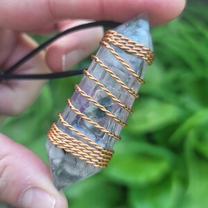 May include: A clear crystal pendant with purple and blue hues, wrapped in gold-colored wire. The crystal is suspended from a black cord. The pendant is held against a backdrop of green foliage.