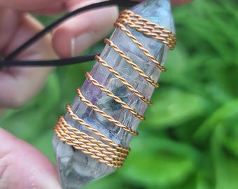 May include: A clear crystal pendant with purple and blue hues, wrapped in gold-colored wire. The crystal is suspended from a black cord. The pendant is held against a backdrop of green foliage.