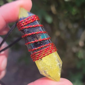 May include: A yellow and black crystal pendant wrapped with red wire, suspended from a black cord. The crystal has a pointed shape and is held against a blurred green background.