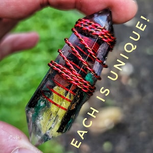 May include: A clear crystal with internal inclusions, wrapped with red and gold wire. The crystal contains green, yellow, and red elements. Text on the side reads "EACH IS UNIQUE!"