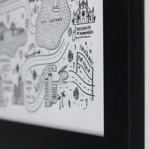 K&ouml;nnte beinhalten: Ein schwarz gerahmter Druck auf wei&szlig;em Hintergrund mit schwarzen Strichzeichnungen von Geb&auml;uden, einem See und Stra&szlig;en. Der Text enth&auml;lt "RECUERDITO DE MEMMINGEN" und "Y MUCHO M&Aacute;S POR VENIR". Das Kunstwerk befindet sich in einem schlichten, d&uuml;nnen schwarzen Rahmen.