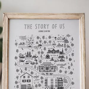 K&ouml;nnte beinhalten: Ein gerahmter Druck mit dem Titel THE STORY OF US, der eine schwarz-wei&szlig;e illustrierte Landkarte mit verschiedenen Sehensw&uuml;rdigkeiten, B&auml;umen und gewundenen Pfaden zeigt. Der Text enth&auml;lt GORINNE & ANTOINE sowie Ortsnamen wie Korsika, &Auml;gypten, Malaysia und Wien.