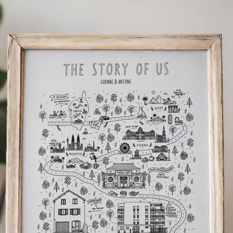 K&ouml;nnte beinhalten: Ein gerahmter Druck mit dem Titel THE STORY OF US, der eine schwarz-wei&szlig;e illustrierte Landkarte mit verschiedenen Sehensw&uuml;rdigkeiten, B&auml;umen und gewundenen Pfaden zeigt. Der Text enth&auml;lt GORINNE & ANTOINE sowie Ortsnamen wie Korsika, &Auml;gypten, Malaysia und Wien.