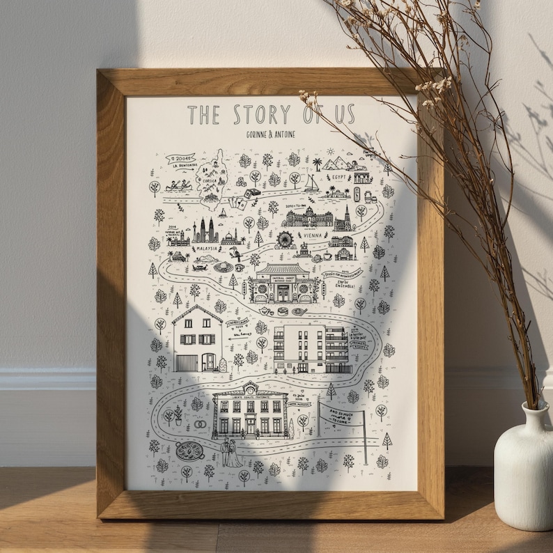 K&ouml;nnte beinhalten: Ein gerahmter Druck mit dem Titel The Story of Us, der eine schwarz-wei&szlig;e illustrierte Karte mit gewundenen Pfaden, verschiedenen Geb&auml;uden, Wahrzeichen und B&auml;umen zeigt. Der Text enth&auml;lt Corinne & Antoine, Malaysia, Vienna und Egypt, in einem Holzrahmen.