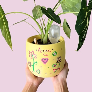 May include: A yellow ceramic plant pot with hand-drawn floral designs and the name "Anna" in pink. The pot holds a green plant with large leaves and a clear plastic watering device. The pot is held by two hands.