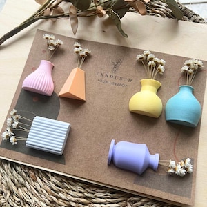 Könnte beinhalten: Eine Sammlung kleiner, 3D-gedruckter Vasen in Pastellfarben wie Rosa, Orange, Gelb, Blau und Lila, jede mit getrockneten weißen Blumen. Die Vasen haben unterschiedliche Formen. Der Text "YANDUS3D YOUR LOVEPOT" ist zu sehen.