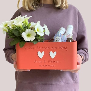 Op de afbeelding: Een koraalkleurige rechthoekige plantenbak met witte bloemen en twee kleine flesjes. De bak heeft de tekst "Zwei Herzen ein Weg", twee witte harten en de namen "Anna & Fabian" met de datum "06.08.2025".