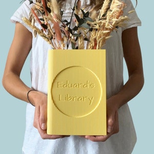 Puede incluir: Una caja amarilla con forma de libro con el texto "Eduard's Library" en relieve en la parte delantera. La caja está siendo sostenida por una persona.