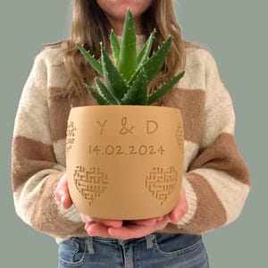 May include: A brown ceramic planter with a heart design and the text "Y & D 14.02.2024" engraved on the front. The planter is holding a green succulent plant.