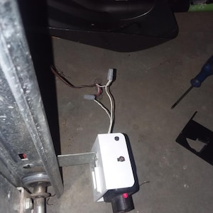 Op de afbeelding: Een witte garagedeuropener sensor met een zwarte basis en rode knop. De sensor is gemonteerd op een metalen beugel en verbonden met draden. Een blauwe schroevendraaier en een zwart plastic onderdeel zijn ook zichtbaar.