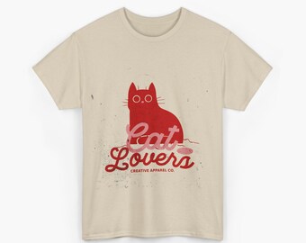 Puede incluir: Una camiseta beige con una ilustración de gato rojo y las palabras "Cat Lovers" en fuente cursiva. La camiseta también incluye el texto "CREATIVE APPAREL CO." debajo del diseño principal.