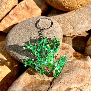 May include: A green frog-shaped keychain with glitter and leaf details. The frog has a silver keyring and a small pink flower on its belly. The keychain is resting on a collection of brown and tan rocks.
