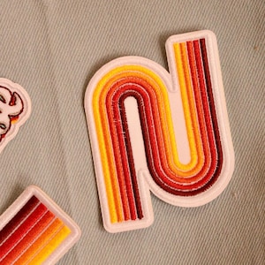 May include: A colourful embroidered patch in a retro style, featuring the letter "N". The design uses a rainbow of colours, including yellow, orange, and red. The patch has a white border and is set against a light blue fabric background.