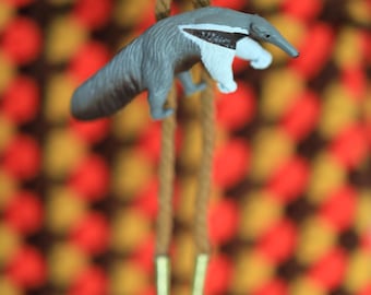 May include: A gray and white anteater figurine hanging from a brown rope with gold tips. The anteater is posed as if it is hanging from the rope.