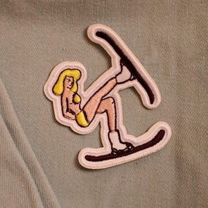 May include: Embroidered patch depicting a cartoon woman in a yellow bikini and blonde hair water skiing. The patch has a white border with brown accents, set against a light blue background.