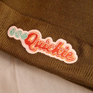 May include: A dark olive green knit beanie with a colorful embroidered patch. The patch features the words "The Quickie" in red and orange lettering with a white border.