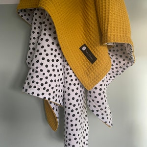 Op de afbeelding: Een babydeken van mosterdgele wafelstof met een wit en zwart stippenpatroon aan de achterkant. De deken heeft een klein label met de tekst "Dotty & Dottie".