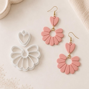 Puede incluir: Un par de pendientes colgantes de color rosa empolvado con una pieza superior en forma de corazón conectada a una pieza inferior en forma de abanico semicircular con ganchos dorados, junto a un cortador de plástico blanco a juego.