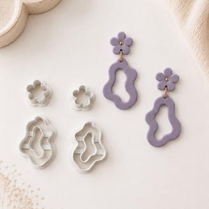 Può includere: Due orecchini pendenti color lavanda composti da un piccolo perno a forma di fiore a cinque petali collegato a una forma aperta irregolare e ondulata. Accanto, quattro stampini in plastica bianca con forme floreali e ondulate.