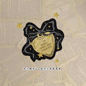 May include: A gold-colored heart-shaped vinyl sticker with the text "I LOVE BADASS FEMALE MAIN CHARACTERS" in black. The sticker has a black border with stars and a bow. The word "VINYL STICKER" is spelled out in white beads at the bottom.