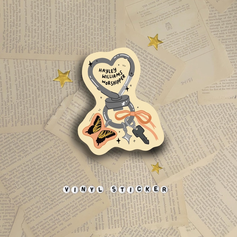 May include: A vinyl sticker featuring a heart-shaped design with a carabiner, keys, and a butterfly illustration. The text "HAYLEY WILLIAMS WORSHIPPER" is written on the heart. The sticker is on a background of vintage book pages, with gold star accents.