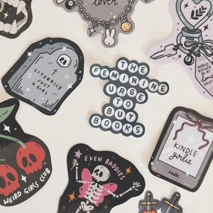May include: A collection of stickers with various designs and text. Designs include a tombstone, a skeleton with butterfly wings, a heart pierced by swords, and the text "The Feminine Urge to Buy Books."