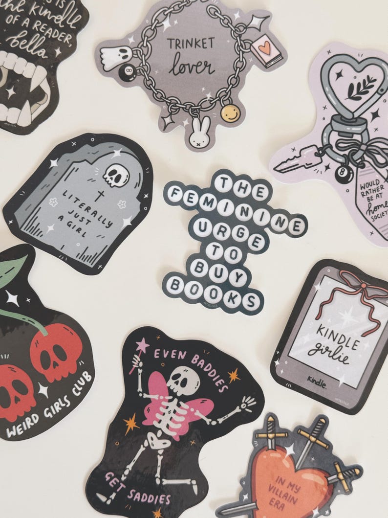 May include: A collection of stickers with various designs and text. Designs include a tombstone, a skeleton with butterfly wings, a heart pierced by swords, and the text "The Feminine Urge to Buy Books."