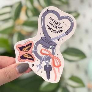 May include: A sticker with a heart-shaped carabiner design, featuring the text "HAYLEY WILLIAMS WORSHIPPER." It includes butterfly illustrations, a key, and a star charm, all in a pastel color palette.