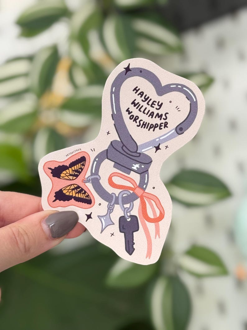 May include: A sticker with a heart-shaped carabiner design, featuring the text "HAYLEY WILLIAMS WORSHIPPER." It includes butterfly illustrations, a key, and a star charm, all in a pastel color palette.