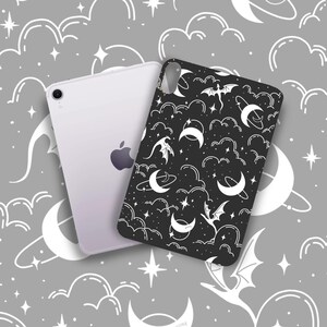May include: A black phone case with a white celestial pattern featuring moons, stars, clouds, and bats. The case is next to a white tablet with the Apple logo. The background is a gray pattern with celestial elements.