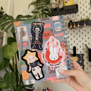 May include: A spiral-bound notebook with a patterned cover featuring various stickers. Stickers include a skeleton, a sun with a face, and a bunny with the text "RAD WOMEN". A yellow cup with "Self Love" text is visible.
