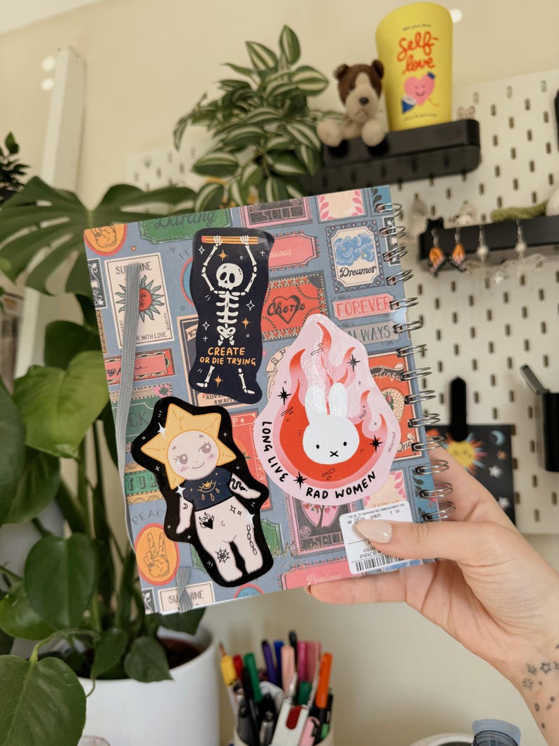 May include: A spiral-bound notebook with a patterned cover featuring various stickers. Stickers include a skeleton, a sun with a face, and a bunny with the text "RAD WOMEN". A yellow cup with "Self Love" text is visible.