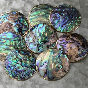 Peut inclure: Plusieurs pendentifs ronds en coquille d'ormeau irisée. Chaque pendentif présente des motifs uniques de bleus, verts et violets, encadrés d'un bord en métal doré. Les pendentifs sont disposés sur une surface grise texturée.