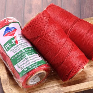 May include: Three spools of red waxed polyester cord, with one still in its packaging. The packaging has text in Portuguese and Spanish. The spools are on a wooden surface.