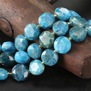 May include: Close-up of a necklace featuring faceted, round blue-green gemstones. The beads are strung together with gold-colored accents. The necklace is resting on a dark wooden surface, showcasing the jewelry's color and texture.